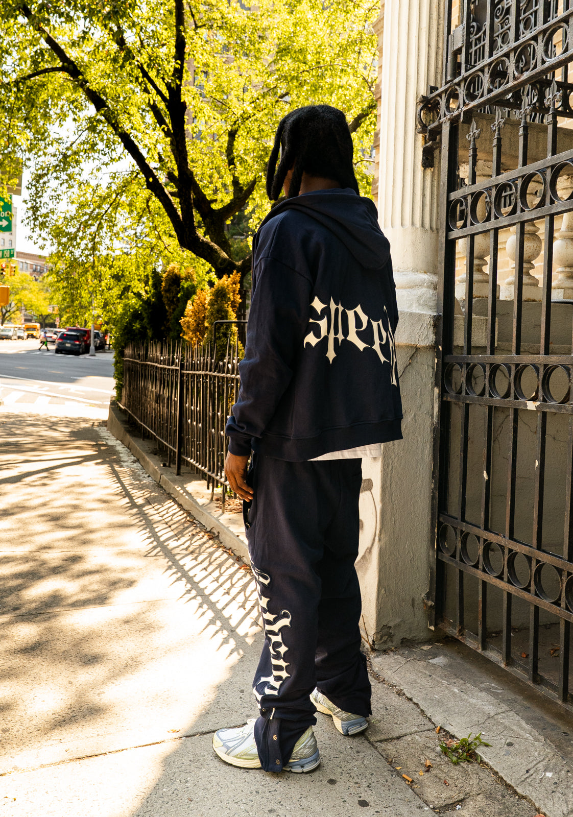 Person wearing a dark hoodie and pants with visible branding, standing near a gated entrance.