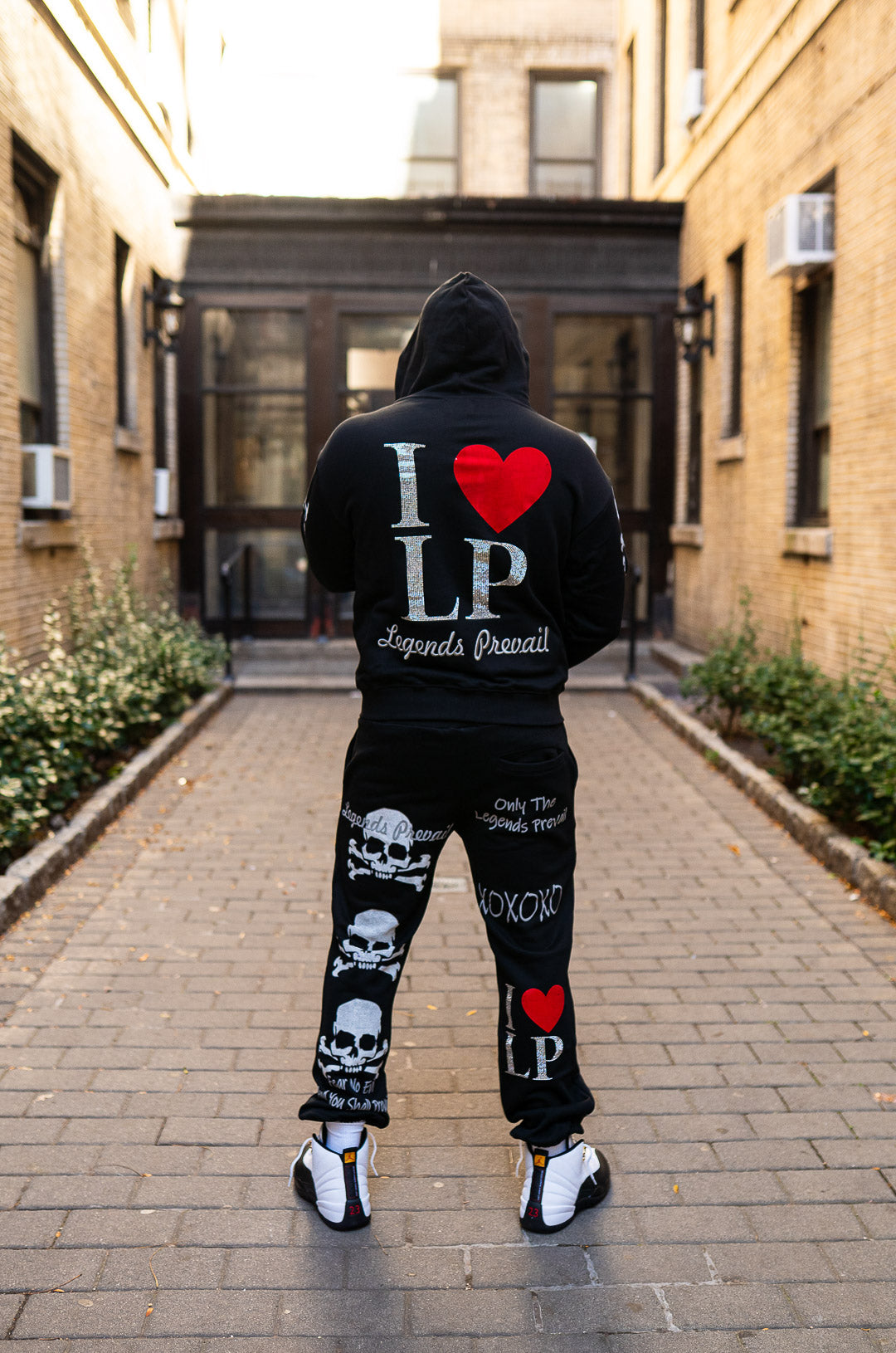 Person wearing a black hoodie and pants with text and heart designs, standing in an alleyway.